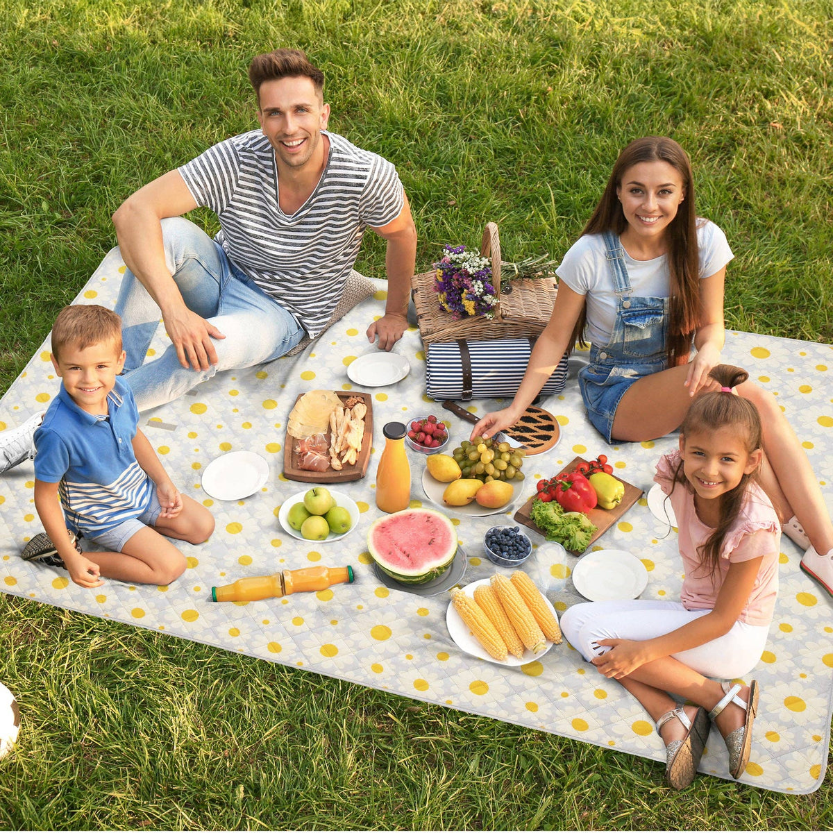 Family of four sitting on yellow daisy-print picnic blanket with food, fresh fruit, and drinks spread across grass