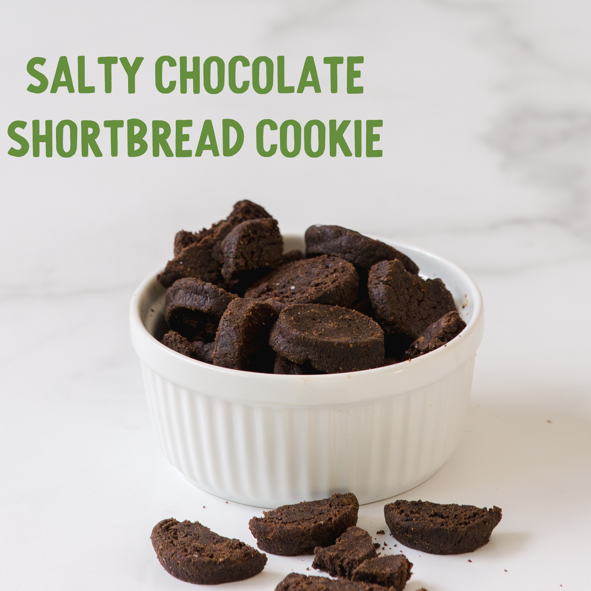 Dark brown salty chocolate shortbread cookies in a white ceramic bowl with scattered pieces on light surface