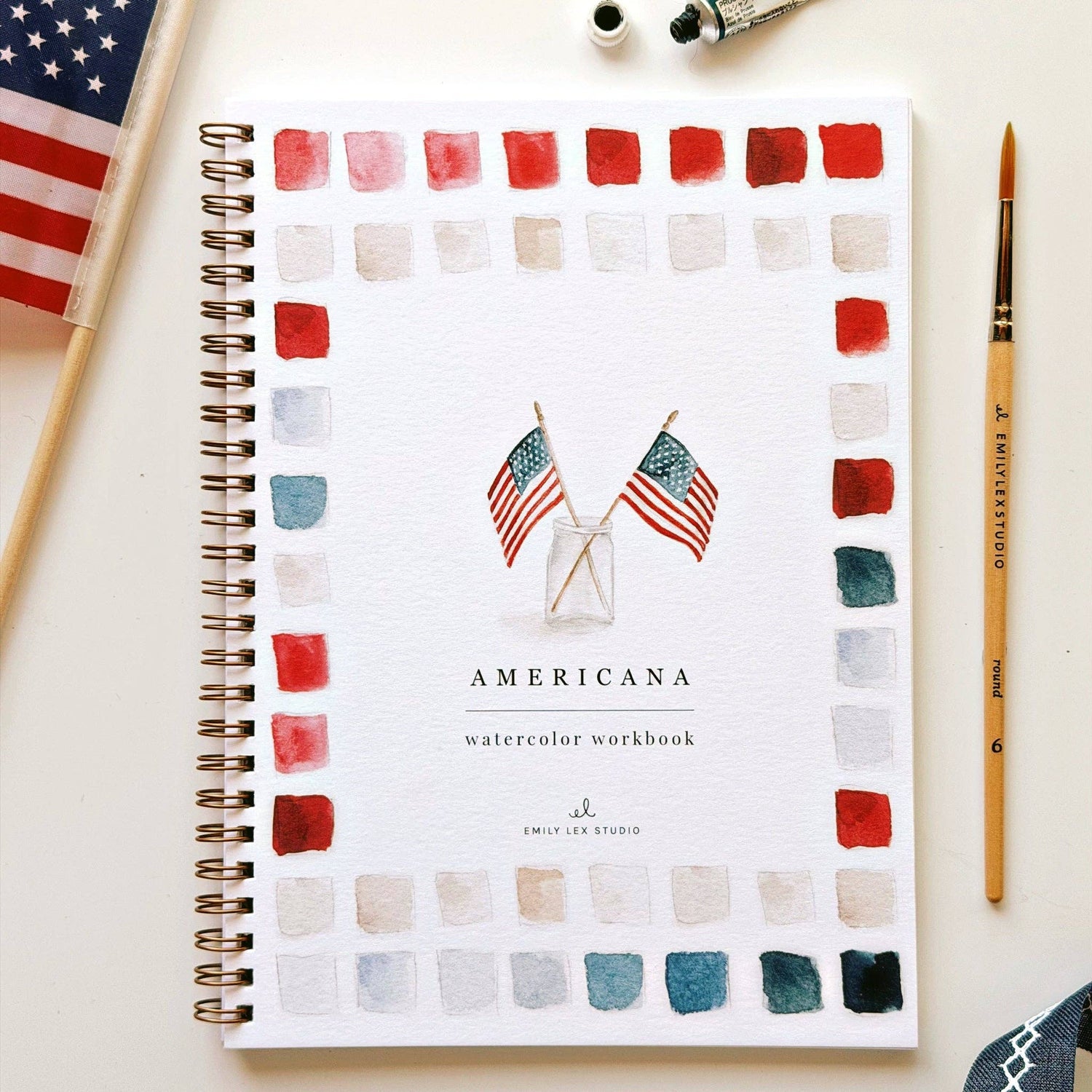 Spiral-bound Americana watercolor workbook with flag illustrations, surrounded by color swatches in reds, pinks, creams, and teals