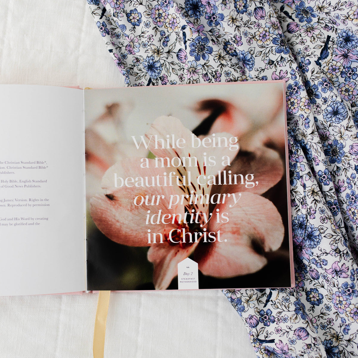 Motherhood Devotional book open on purple floral fabric, cover showing hands holding infant with text overlay: "While being a mom is a beautiful calling, our primary identity is in Christ."