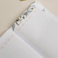 Gardening journal open with botanical watercolor illustrations featuring seeds and leaves, paired with gold-trim pen on neutral background