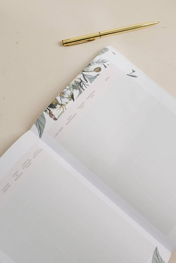 Gardening journal open with botanical watercolor illustrations featuring seeds and leaves, paired with gold-trim pen on neutral background