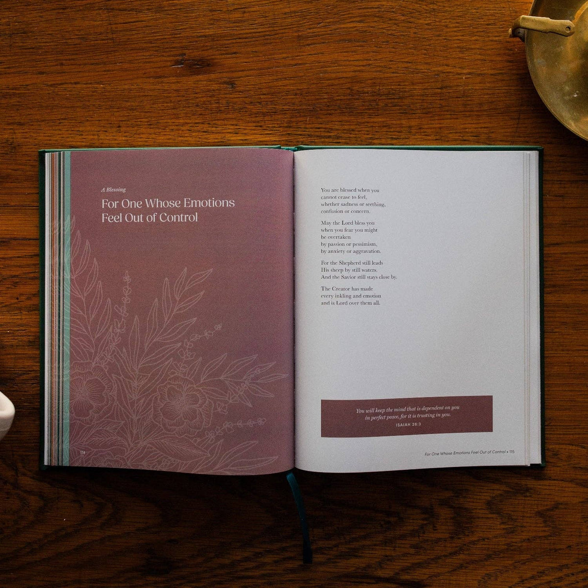 Open prayer book with mauve left page featuring floral design and white text, and white right page with burgundy scripture banner on wooden surface.