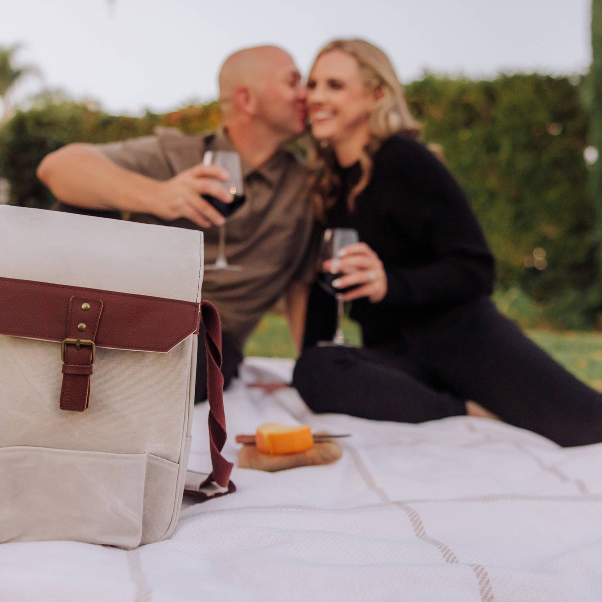 Cream insulated wine tote with burgundy leather flap, displayed on grass during outdoor picnic