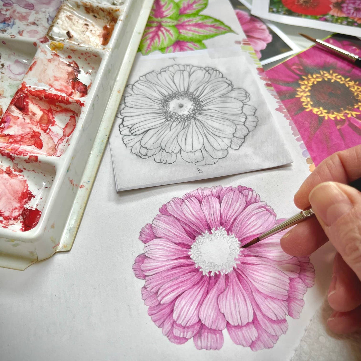 Hand coloring a pink daisy illustration with marker next to an uncolored sketch and colorful floral design papers in a white organizer tray