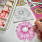 Hand coloring a pink daisy illustration with marker next to an uncolored sketch and colorful floral design papers in a white organizer tray