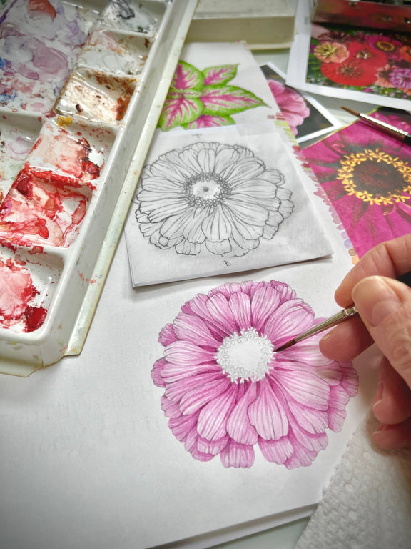 Hand coloring a pink daisy illustration with marker next to an uncolored sketch and colorful floral design papers in a white organizer tray