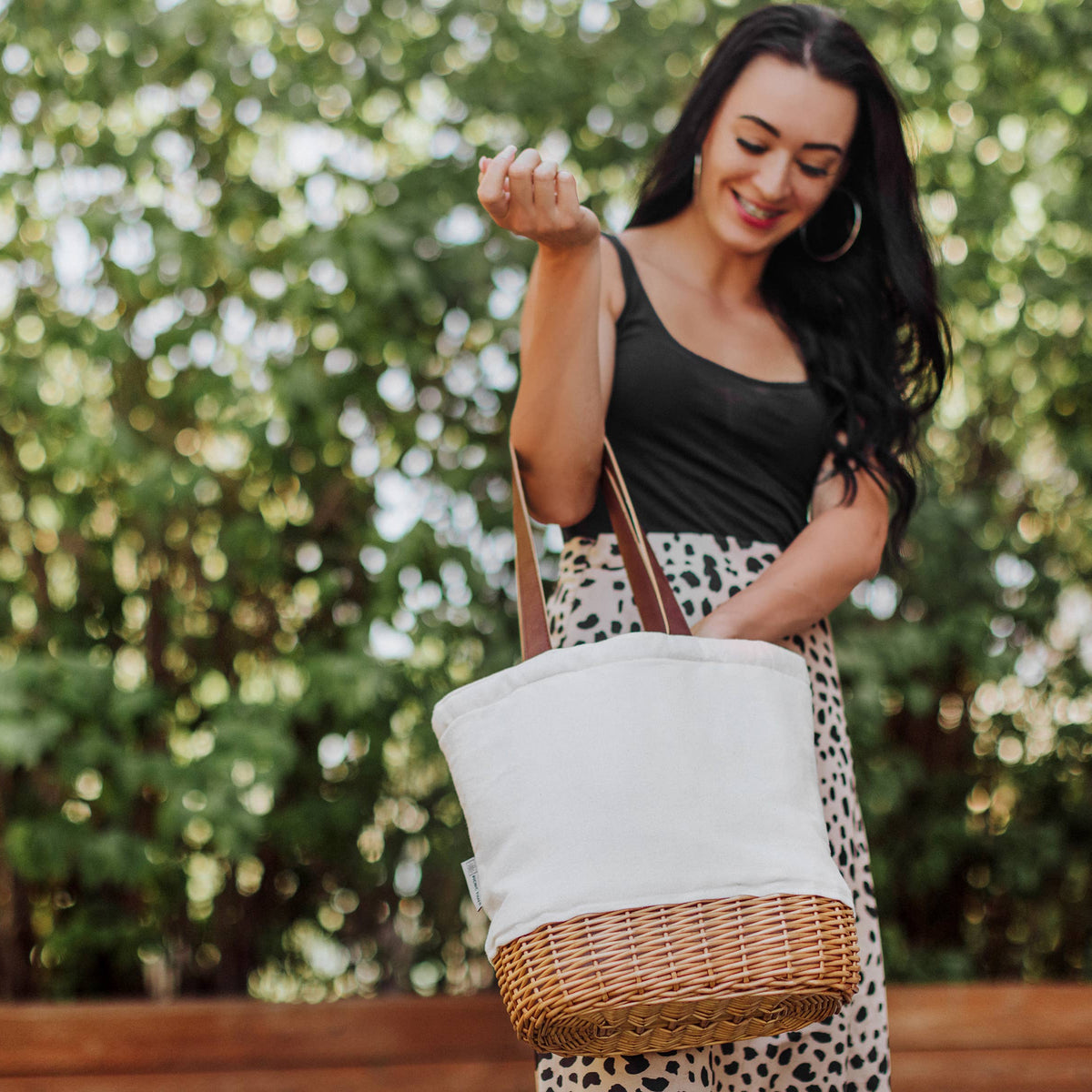Woman wearing black tank top carrying cream canvas tote with natural wicker base and tan leatherette handles in sunlit outdoor setting