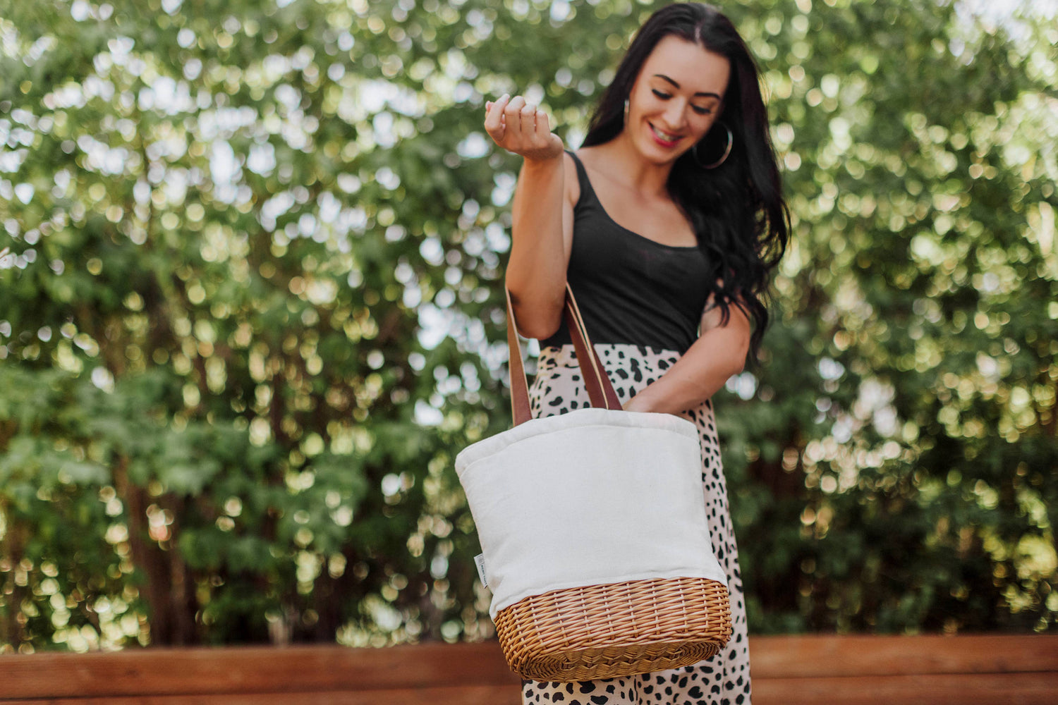 Woman wearing black tank top carrying cream canvas tote with natural wicker base and tan leatherette handles in sunlit outdoor setting