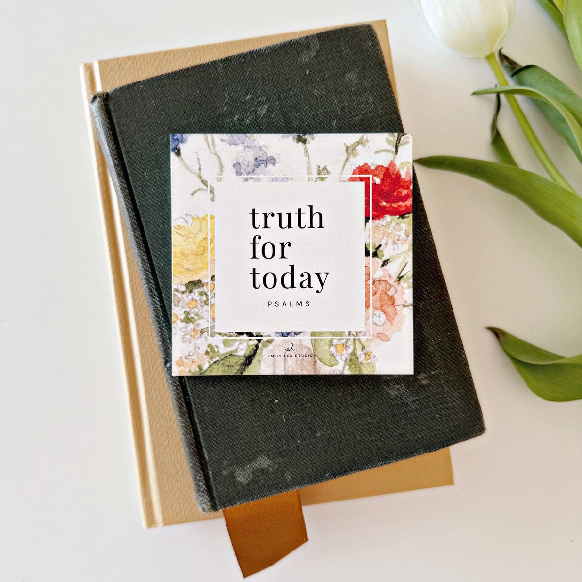A set of square cards with floral watercolor illustrations and text, placed on a stack of books with a tulip flower beside it.