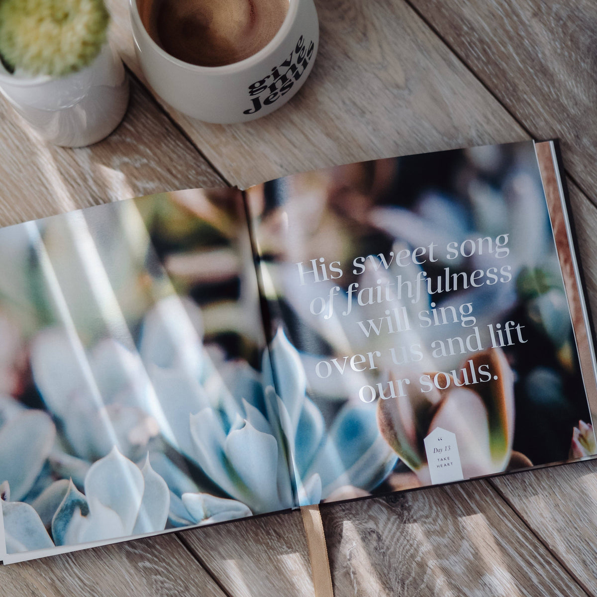 Open devotional book with blue succulent cover and inspirational quote, displayed on wood with potted succulent and ceramic mug.