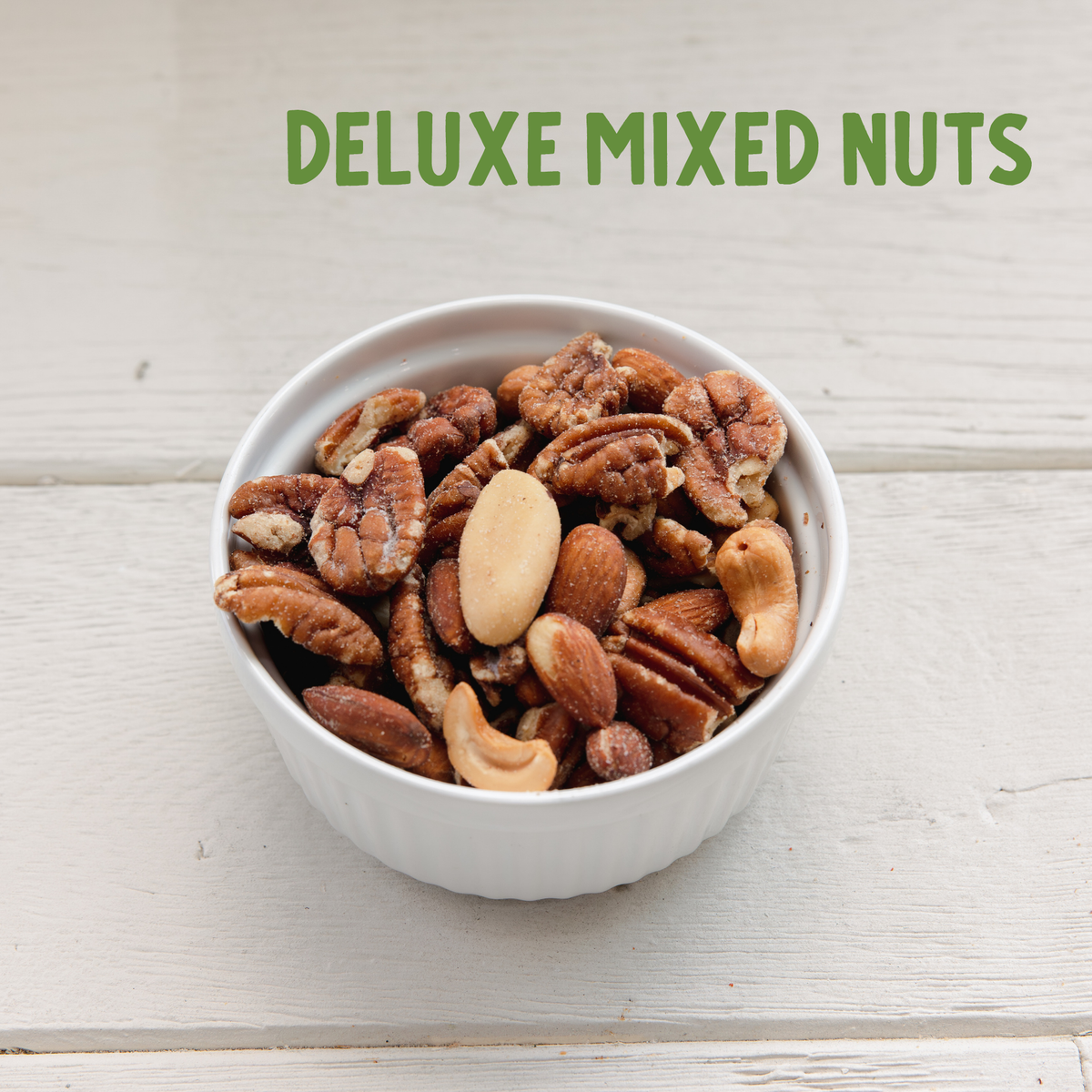 White ceramic bowl overflowing with roasted mixed nuts including cashews, almonds, and pecans on weathered white wood.