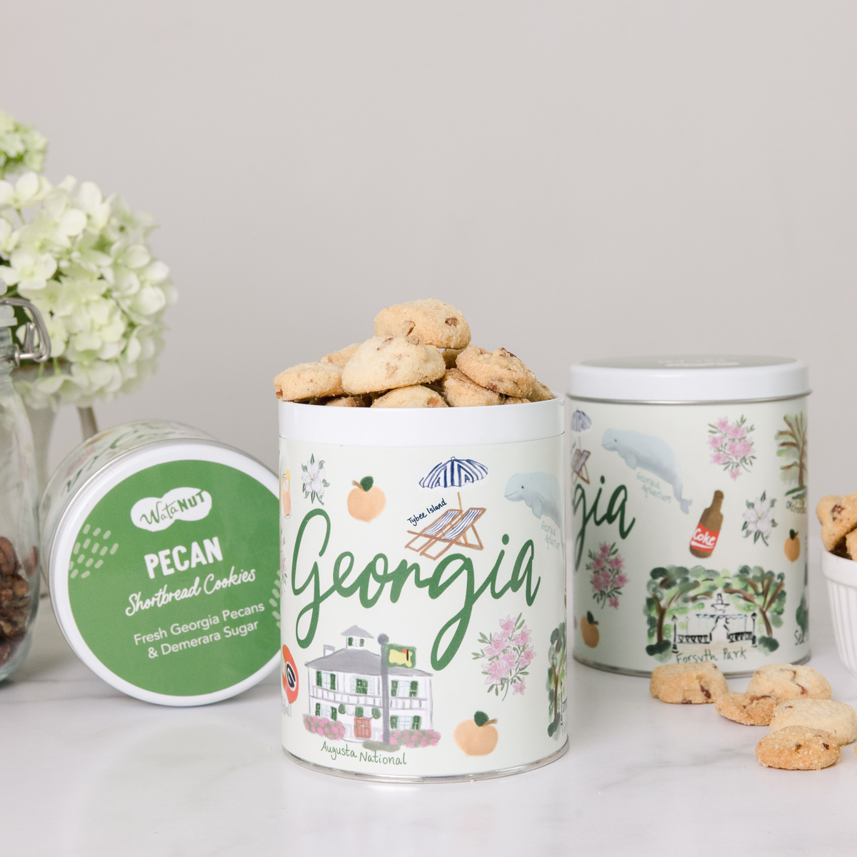 Three white Georgia state-themed gift tins with peach and building motifs, displaying gourmet cookies and crackers alongside fresh white flowers.