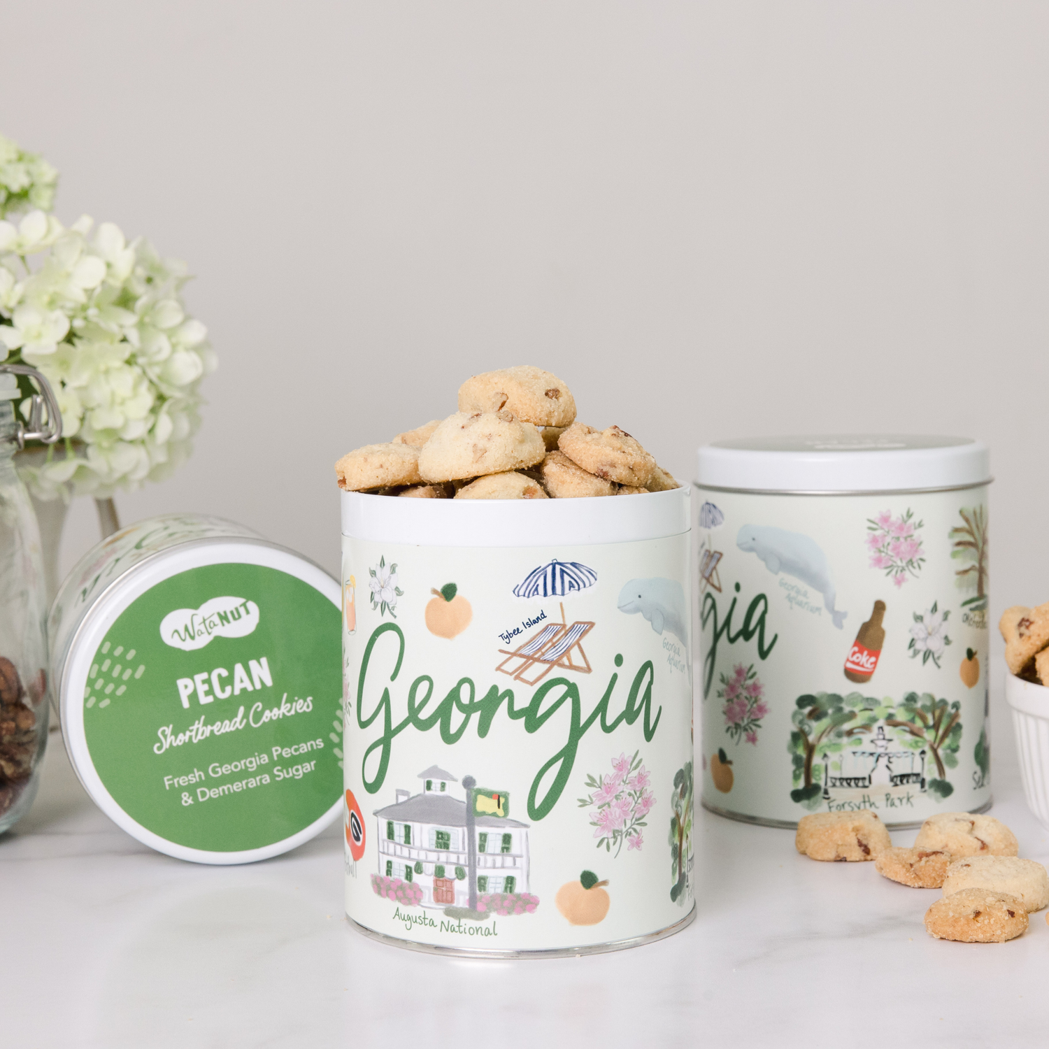 Three white Georgia state-themed gift tins with peach and building motifs, displaying gourmet cookies and crackers alongside fresh white flowers.