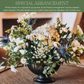 Lush floral arrangement in black pedestal vase with white daisies, cream roses, light blue hydrangeas, delicate baby's breath, thistles, and mixed greenery