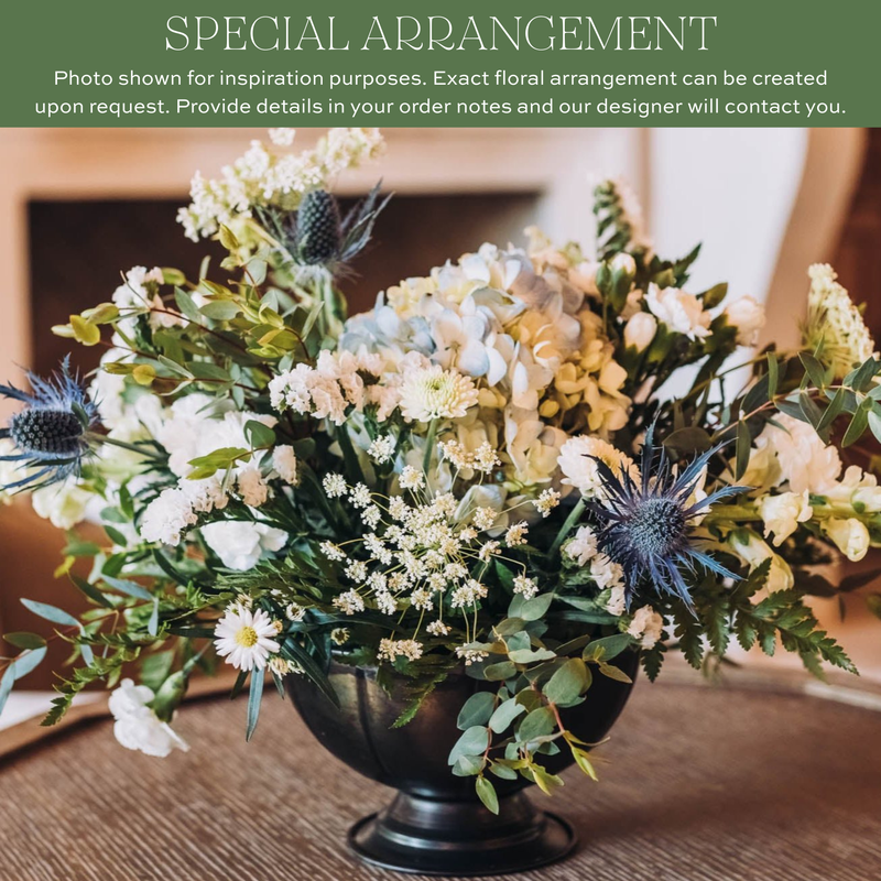 Lush floral arrangement in black pedestal vase with white daisies, cream roses, light blue hydrangeas, delicate baby's breath, thistles, and mixed greenery
