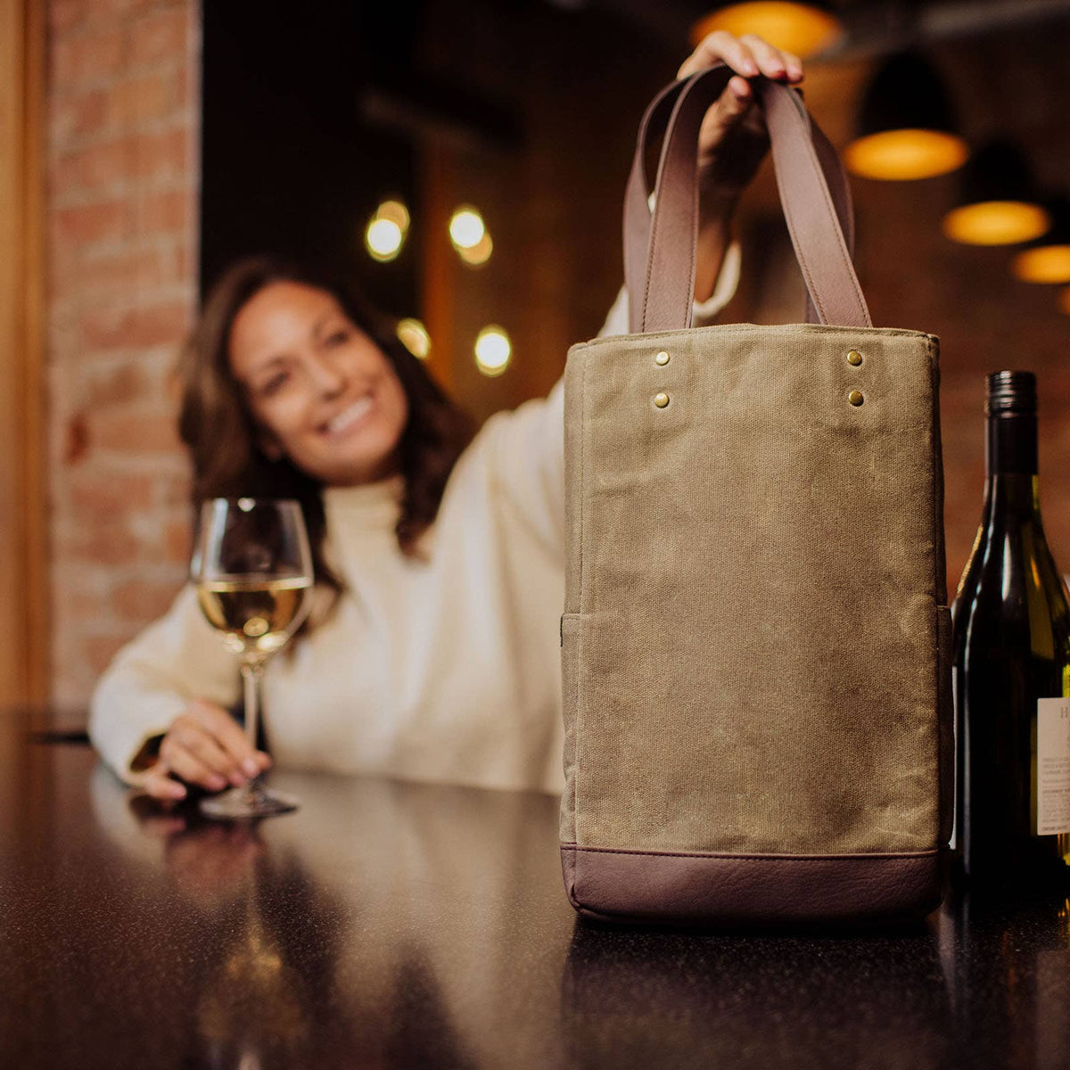 Beige canvas insulated wine tote with brown leather bottom panel and dual handles, holding a wine bottle on a dark table with warm lighting