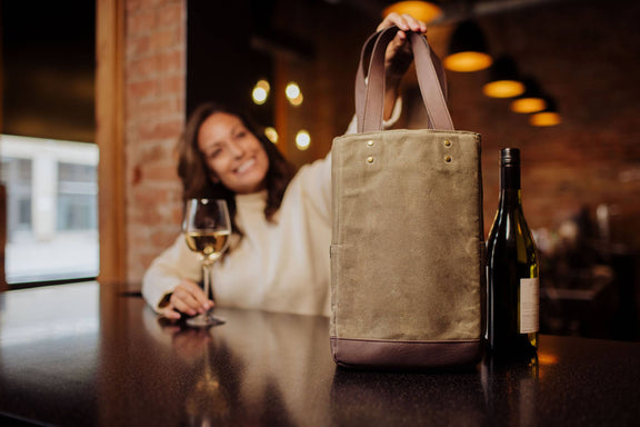 Beige canvas insulated wine tote with brown leather bottom panel and dual handles, holding a wine bottle on a dark table with warm lighting