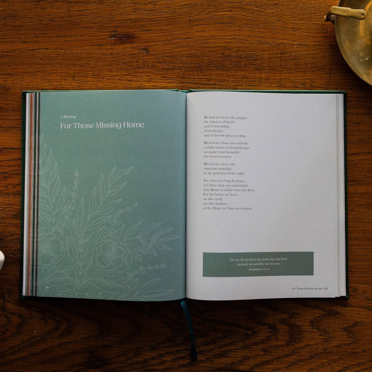 Open book with sage green page featuring botanical leaves and "For Those Missing Home" blessing, alongside prayer text page with green quote box, set on wooden surface.