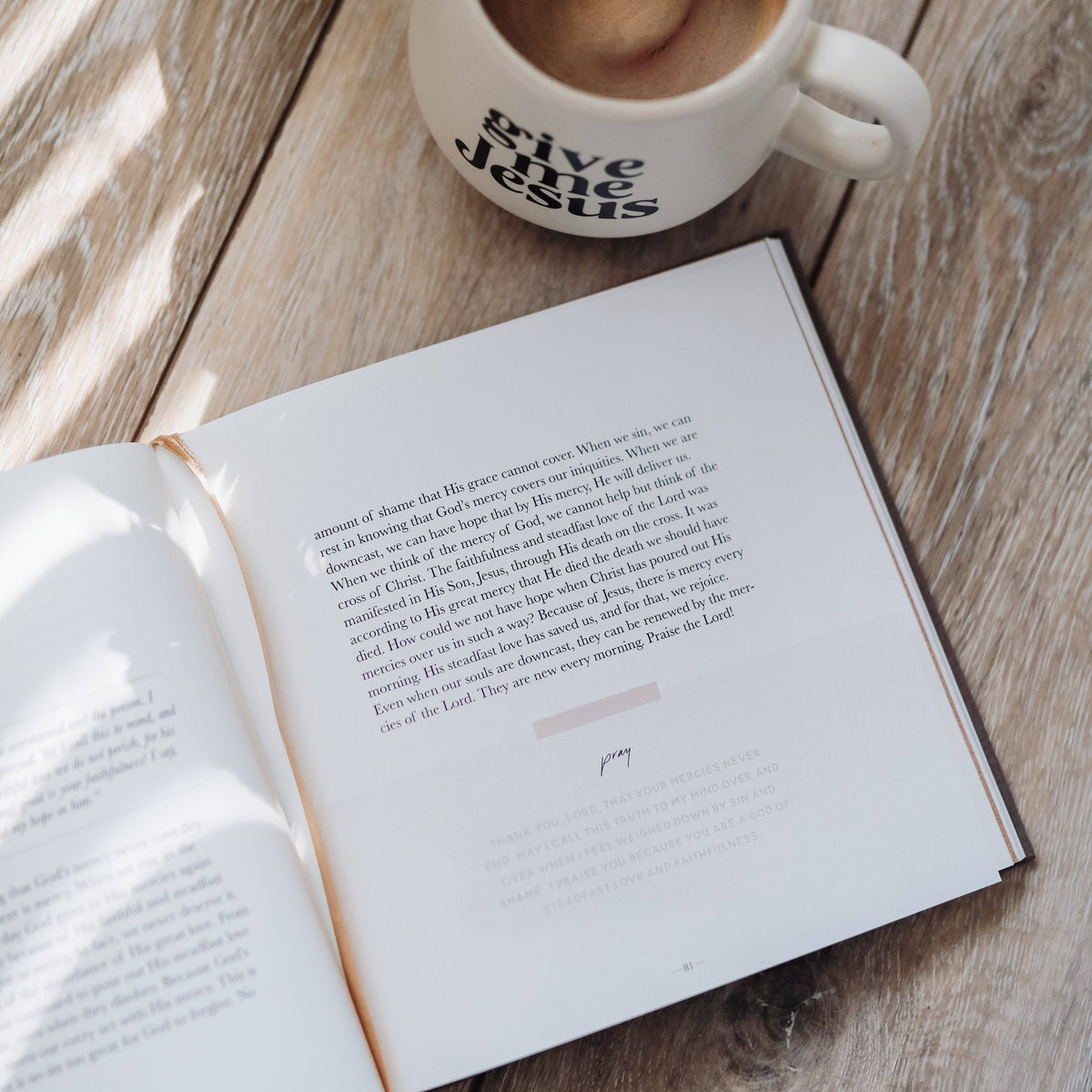 Open devotional book with scripture passage on cream-colored pages, placed on weathered wood with gold bookmark ribbon and ceramic mug above.