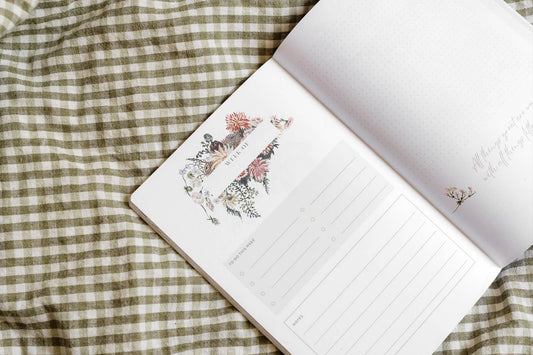 Open gardening journal with botanical flower and plant illustrations on cream pages and lined writing space, displayed on green gingham fabric.