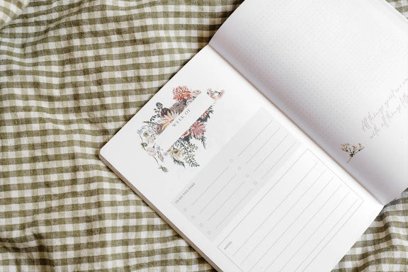 Open gardening journal with botanical flower and plant illustrations on cream pages and lined writing space, displayed on green gingham fabric.