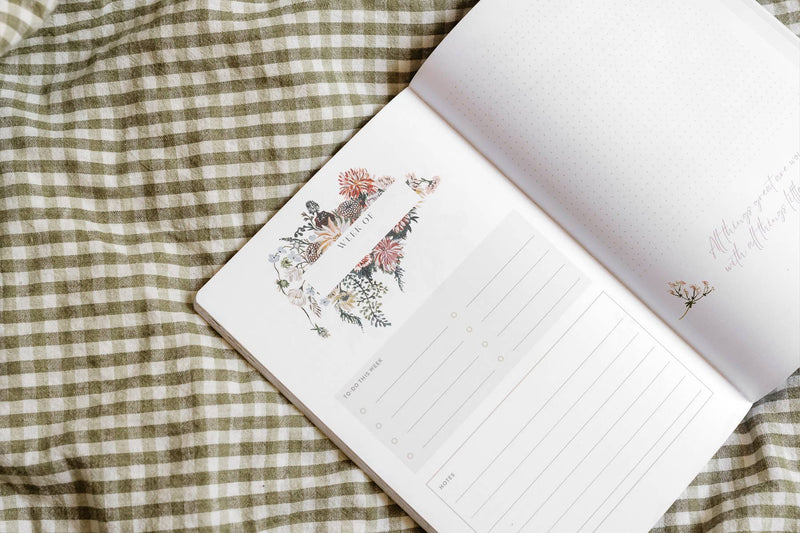 Open gardening journal with botanical flower and plant illustrations on cream pages and lined writing space, displayed on green gingham fabric.