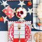Red and white star-patterned paper cups styled in patriotic table setting with navy background, checkered napkins, fresh berries, and gold utensils.