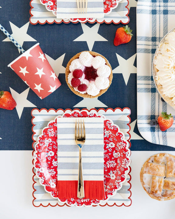 Red and white star-patterned paper cups styled in patriotic table setting with navy background, checkered napkins, fresh berries, and gold utensils.