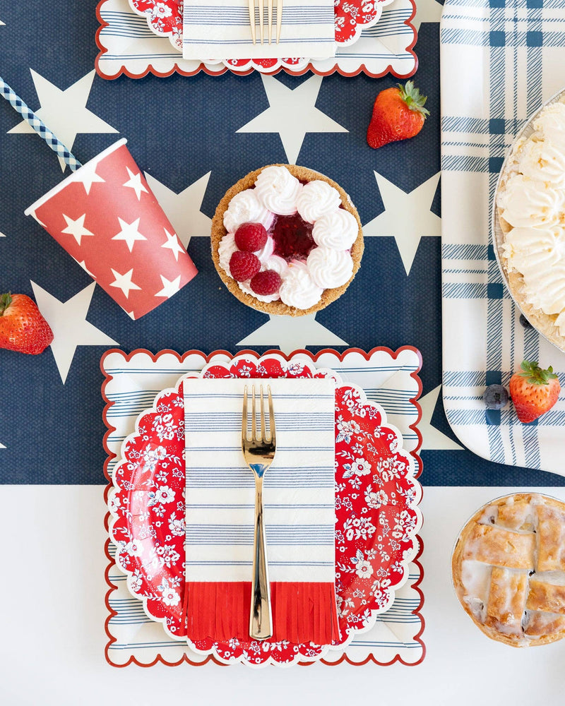 Red and white star-patterned paper cups styled in patriotic table setting with navy background, checkered napkins, fresh berries, and gold utensils.