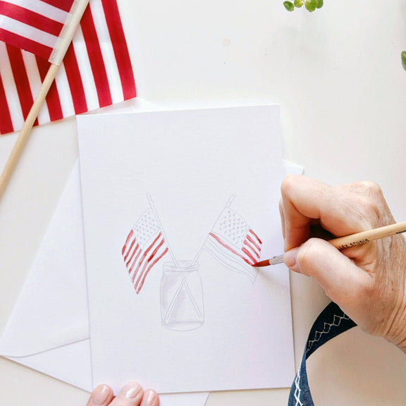 Hand painting red and white striped American flags in a mason jar outline on a white notecard, with a paintbrush mid-stroke and decorative flag nearby
