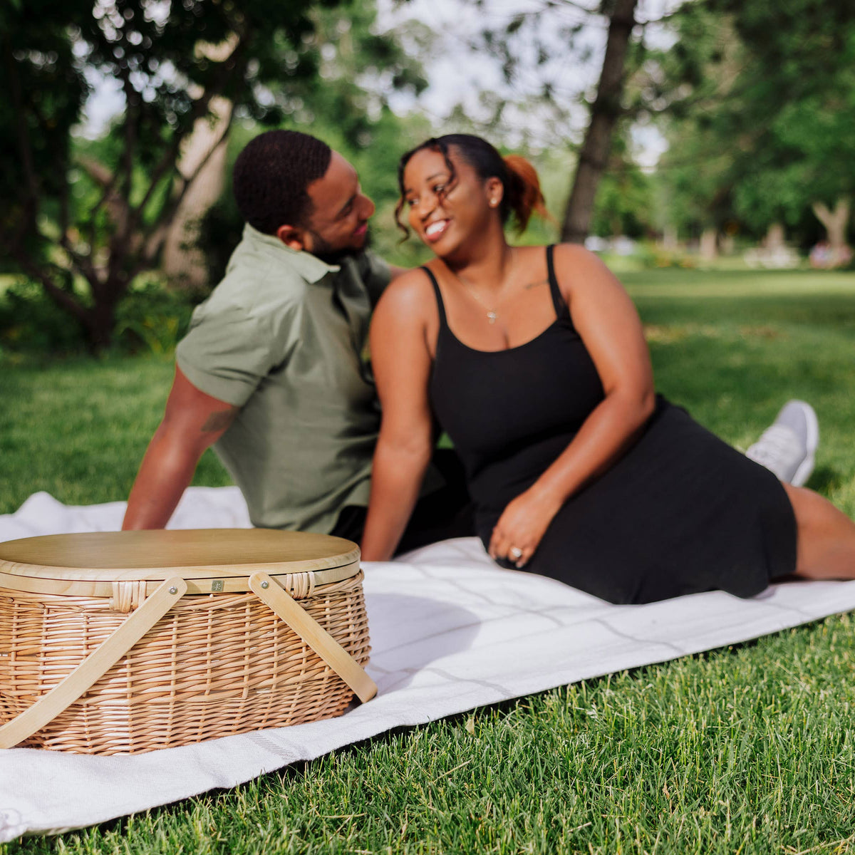 Beige woven wicker picnic basket with canvas straps and lid on outdoor picnic blanket