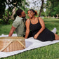 Beige woven wicker picnic basket with canvas straps and lid on outdoor picnic blanket