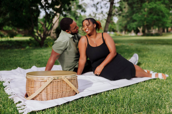 Beige woven wicker picnic basket with canvas straps and lid on outdoor picnic blanket