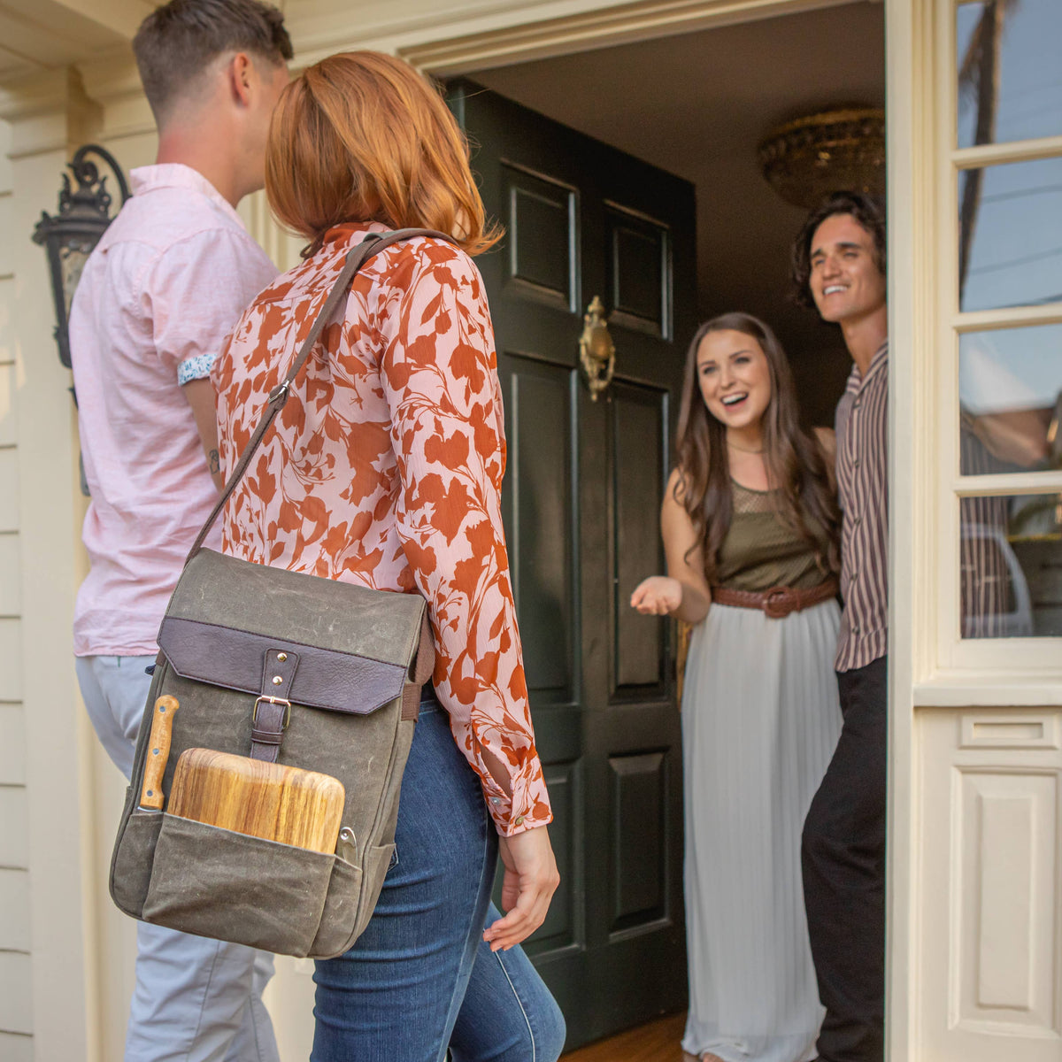 Gray insulated wine backpack with burgundy leather flap and cheese board tucked in front pocket, worn by woman at doorway