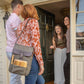 Gray insulated wine backpack with burgundy leather flap and cheese board tucked in front pocket, worn by woman at doorway