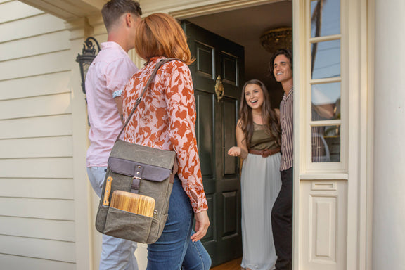 Gray insulated wine backpack with burgundy leather flap and cheese board tucked in front pocket, worn by woman at doorway