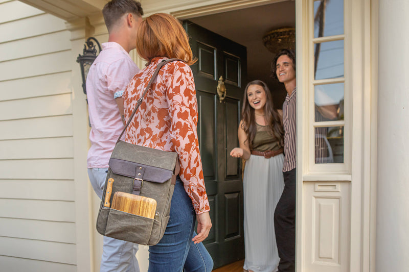 Gray insulated wine backpack with burgundy leather flap and cheese board tucked in front pocket, worn by woman at doorway