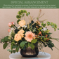 Peach roses and pink gerbera daisies with mixed greenery and ferns in a black rounded vase on neutral background.