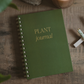 Forest green spiral-bound plant journal with gold foil text, displayed on wooden surface with plant sprigs and pen
