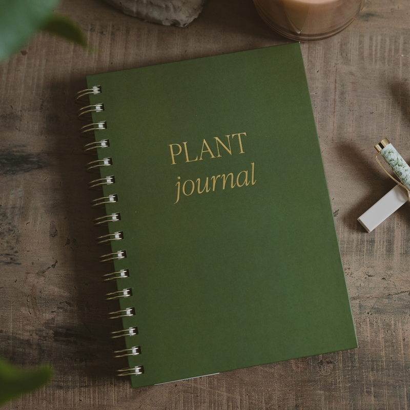 Forest green spiral-bound plant journal with gold foil text, displayed on wooden surface with plant sprigs and pen