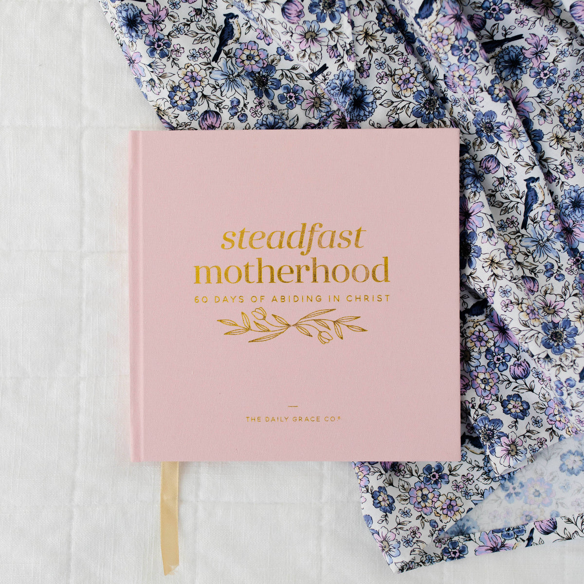 Pink hardcover devotional "Steadfast Motherhood" with gold foil lettering and bookmark, styled with blue floral fabric