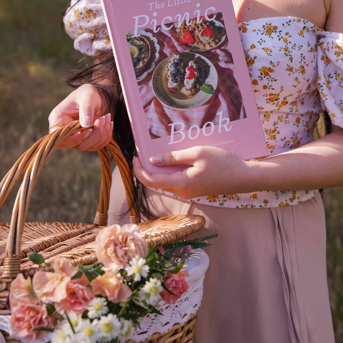 Purple-covered picnic book held by woman in floral top carrying woven basket of pink and white flowers in sunlit outdoor setting