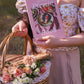 Purple-covered picnic book held by woman in floral top carrying woven basket of pink and white flowers in sunlit outdoor setting