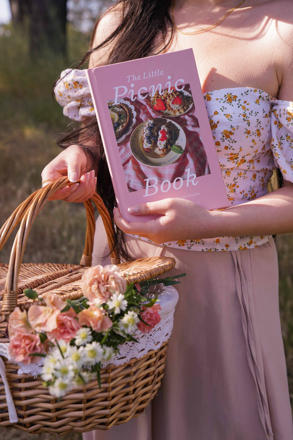 Purple-covered picnic book held by woman in floral top carrying woven basket of pink and white flowers in sunlit outdoor setting