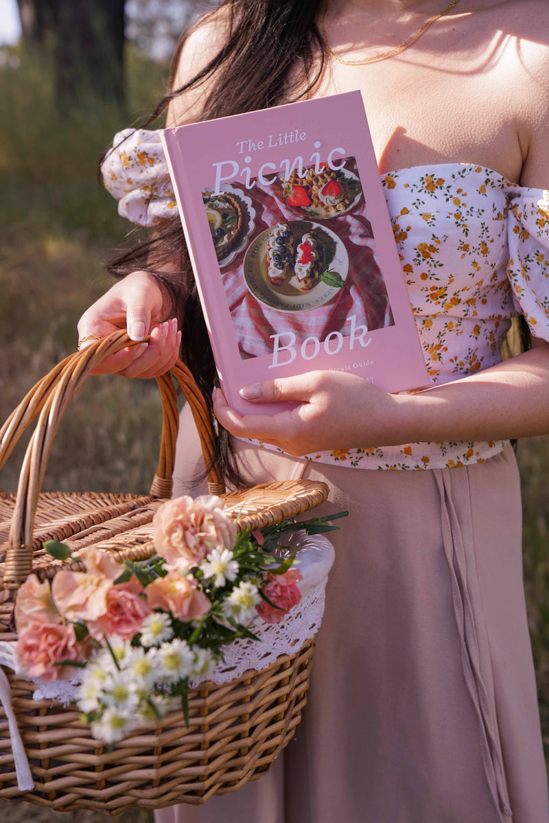 Purple-covered picnic book held by woman in floral top carrying woven basket of pink and white flowers in sunlit outdoor setting