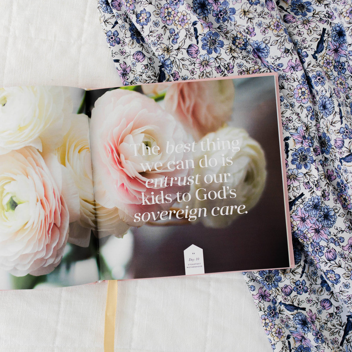 Motherhood Devotional book cover featuring inspirational quote on blurred pink and cream florals, displayed with fresh roses and vintage floral fabric