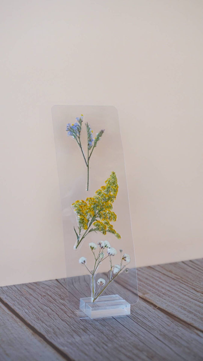 Pressed flower bookmark with blue forget-me-nots, yellow yarrow, and pale blue baby's breath on clear acrylic stand.