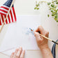 Hand painting white notecard with watercolor brush in shades of blue, styled with American flag and green foliage