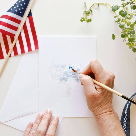 Hand painting white notecard with watercolor brush in shades of blue, styled with American flag and green foliage
